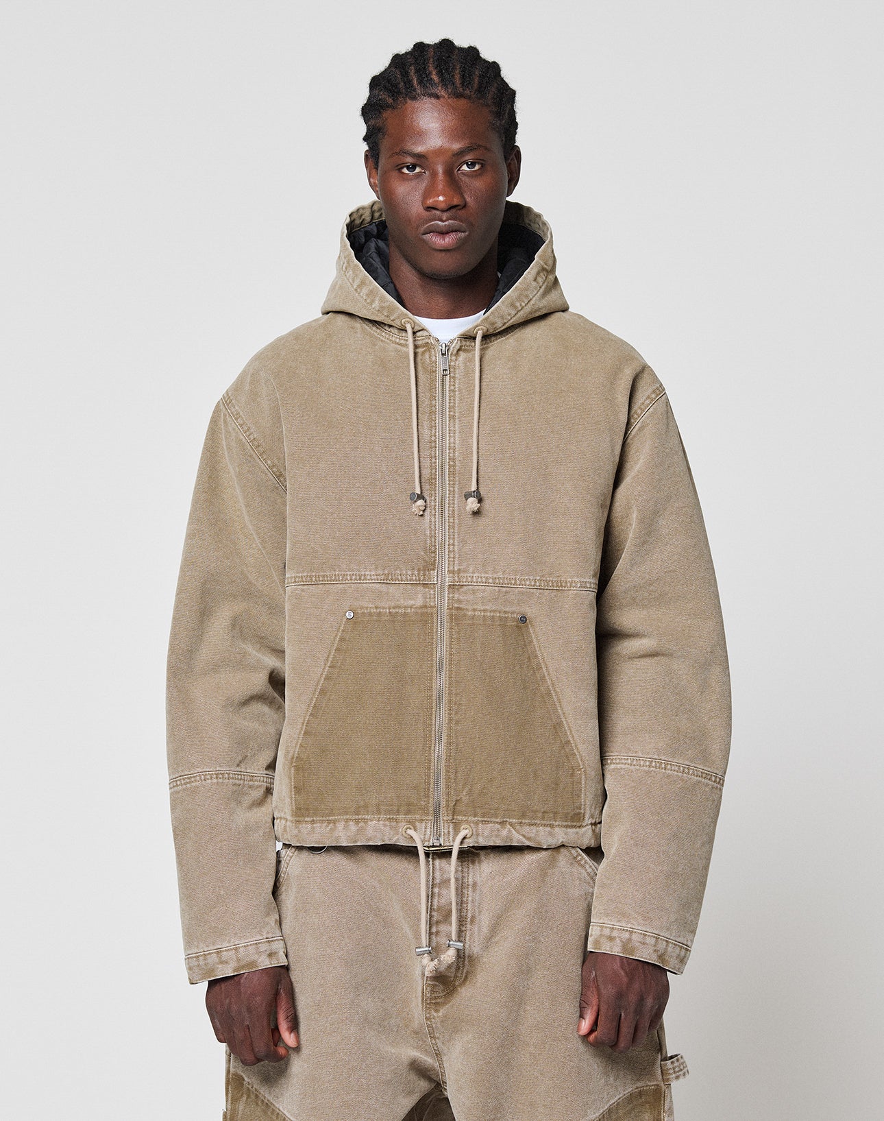 A person with medium-length braided hair wears a tan Faded Workwear Jacket by LIVE FAST DIE YOUNG and matching tan pants, standing against a plain light gray background. | model