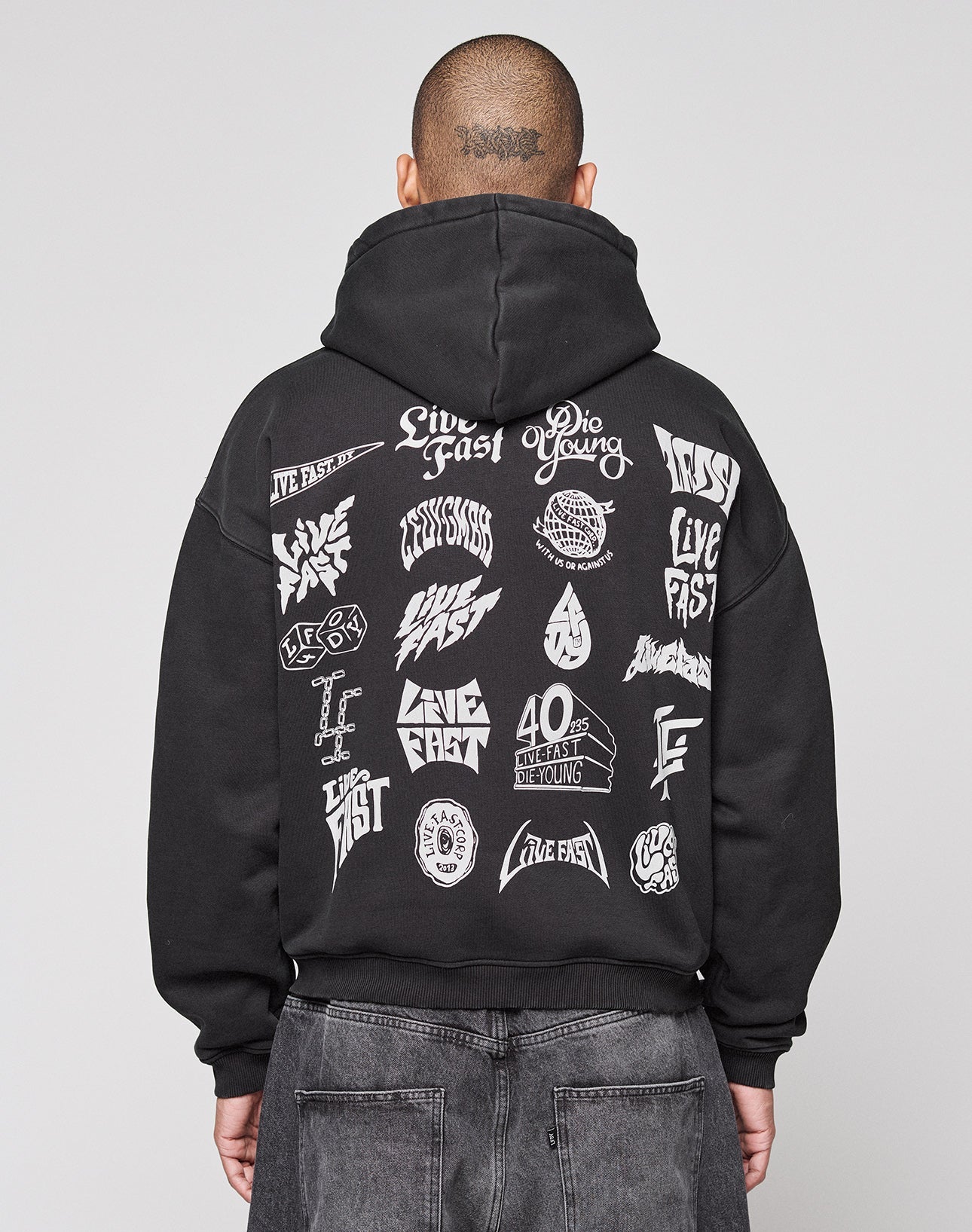 A person faces away from the camera in a black LIVE FAST DIE YOUNG LFDY Logo Hooded Zip, an oversized Baumwolle hoodie with multilogo white graphics and “Live Fast” and “Die Young” phrases on the back, set against a plain gray background. | model