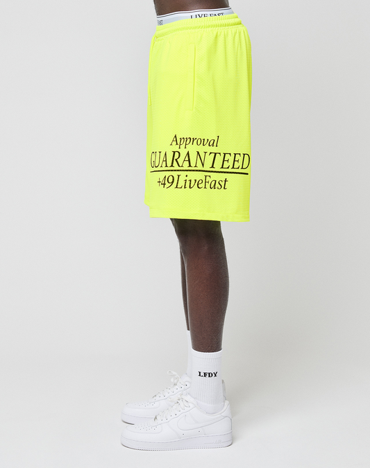 A person stands sideways wearing neon yellow LFDY Guaranteed Mesh Shorts by LIVE FAST DIE YOUNG with black Approval GUARANTEED +49LiveFast text, white LEDX socks, and white Nike sneakers against a plain white background.