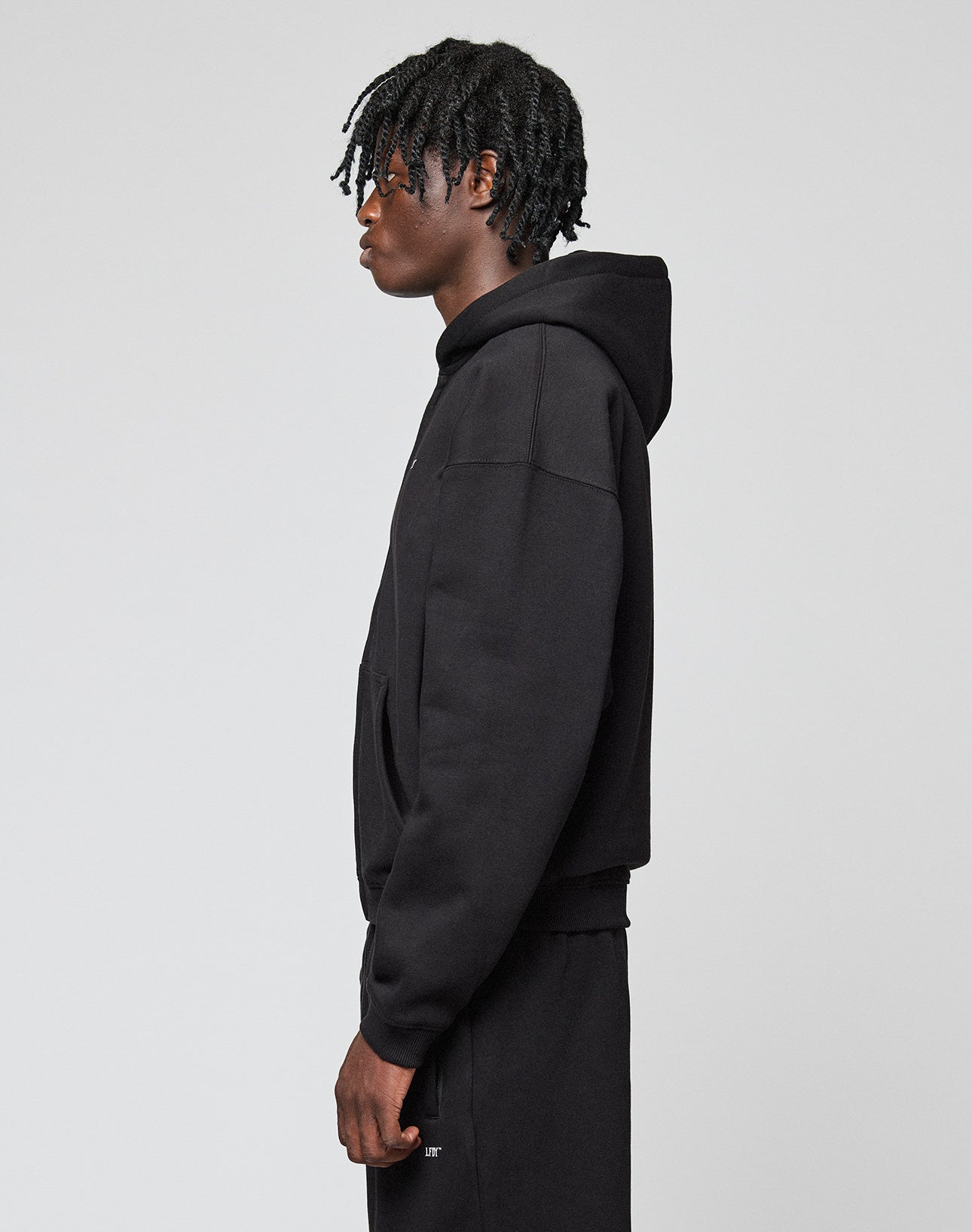 A person stands in profile against a plain light background, wearing the LIVE FAST DIE YOUNG Basic Hooded Zip and matching black pants. Their hair is styled in short twists, and their hands rest relaxed at their sides.