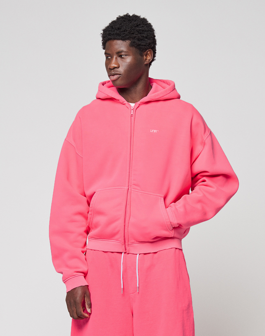 A person in a bright pink Basic Hooded Zip and matching sweatpants from LIVE FAST DIE YOUNG, featuring subtle branding, stands against a plain light gray background, gazing slightly to the side with one hand in their pocket.