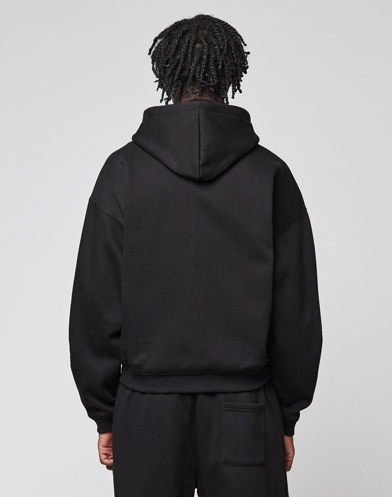 A person with short, twisted hair faces away from the camera, wearing a black LIVE FAST DIE YOUNG Basic Hooded Zip sweatshirt and black pants against a plain light background.
