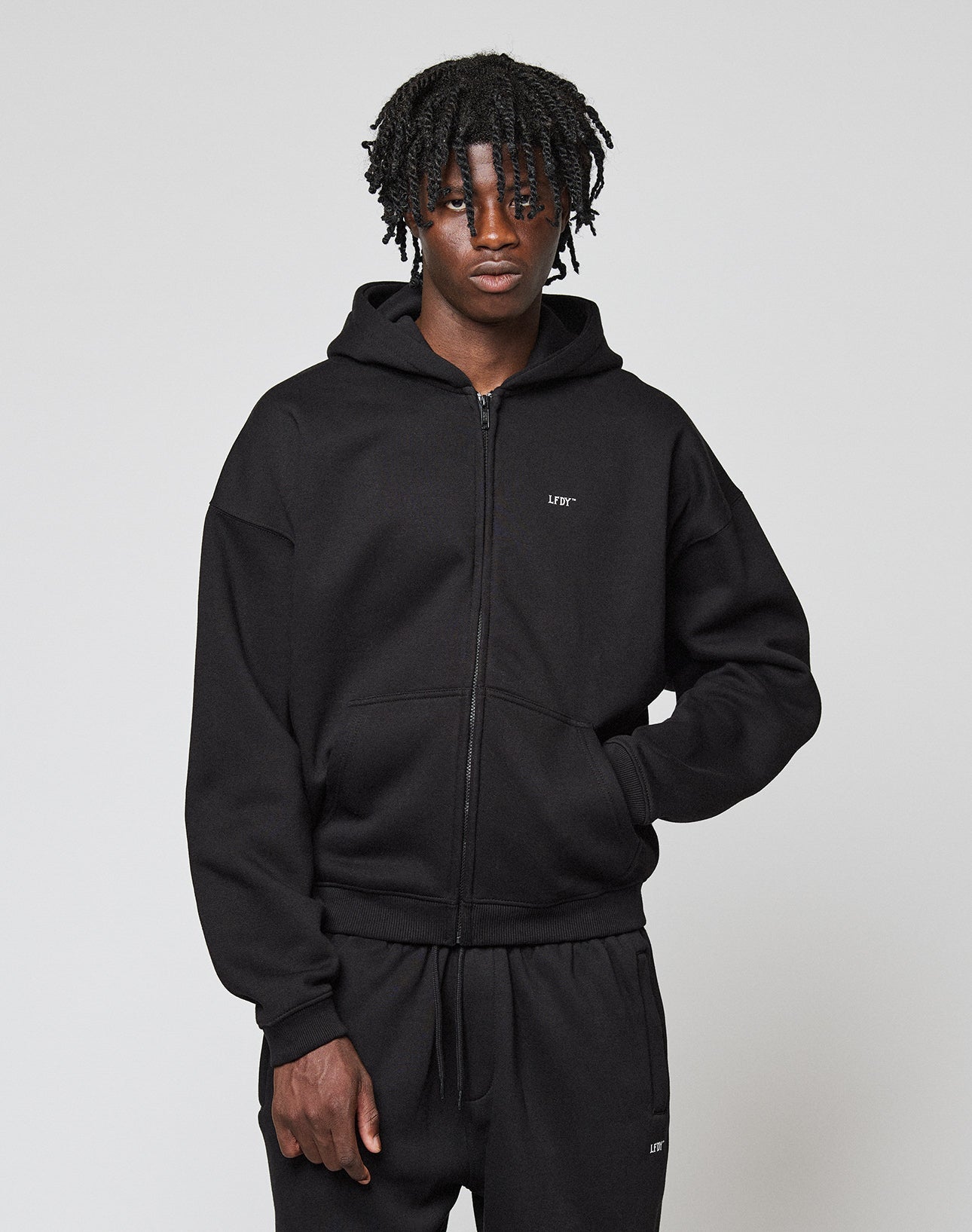 A person with short, curly hair wears the Basic Hooded Zip by LIVE FAST DIE YOUNG and matching black sweatpants, standing against a plain light gray background.