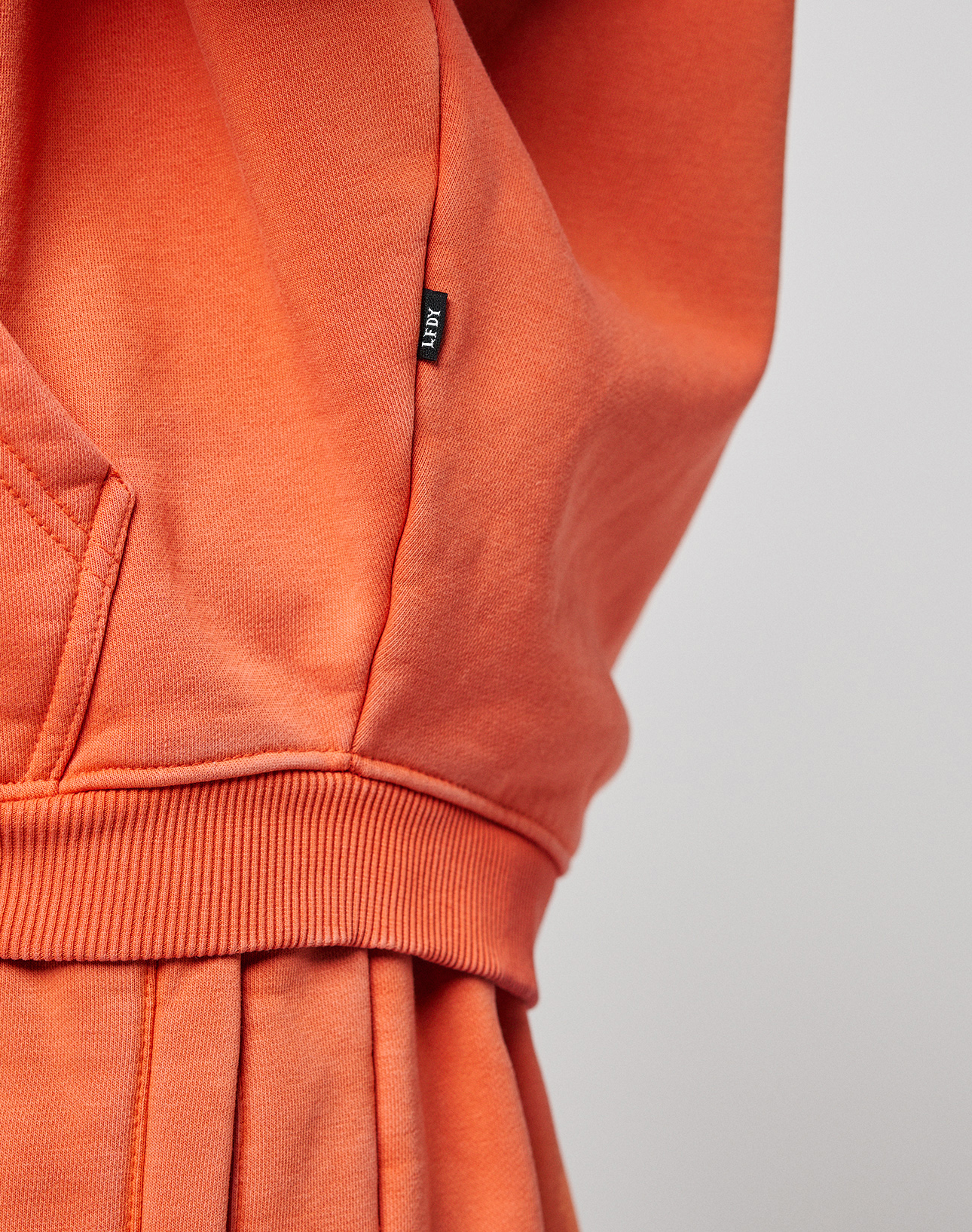 Close-up of an orange LFDY 40 Hooded Zip from LIVE FAST DIE YOUNG, featuring a ribbed waistband, front pocket corner, and small black tag. The plain light background highlights its Oversize Fit for a relaxed look.