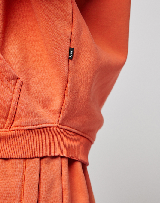 Close-up of an orange LFDY 40 Hooded Zip from LIVE FAST DIE YOUNG, featuring a ribbed waistband, front pocket corner, and small black tag. The plain light background highlights its Oversize Fit for a relaxed look.