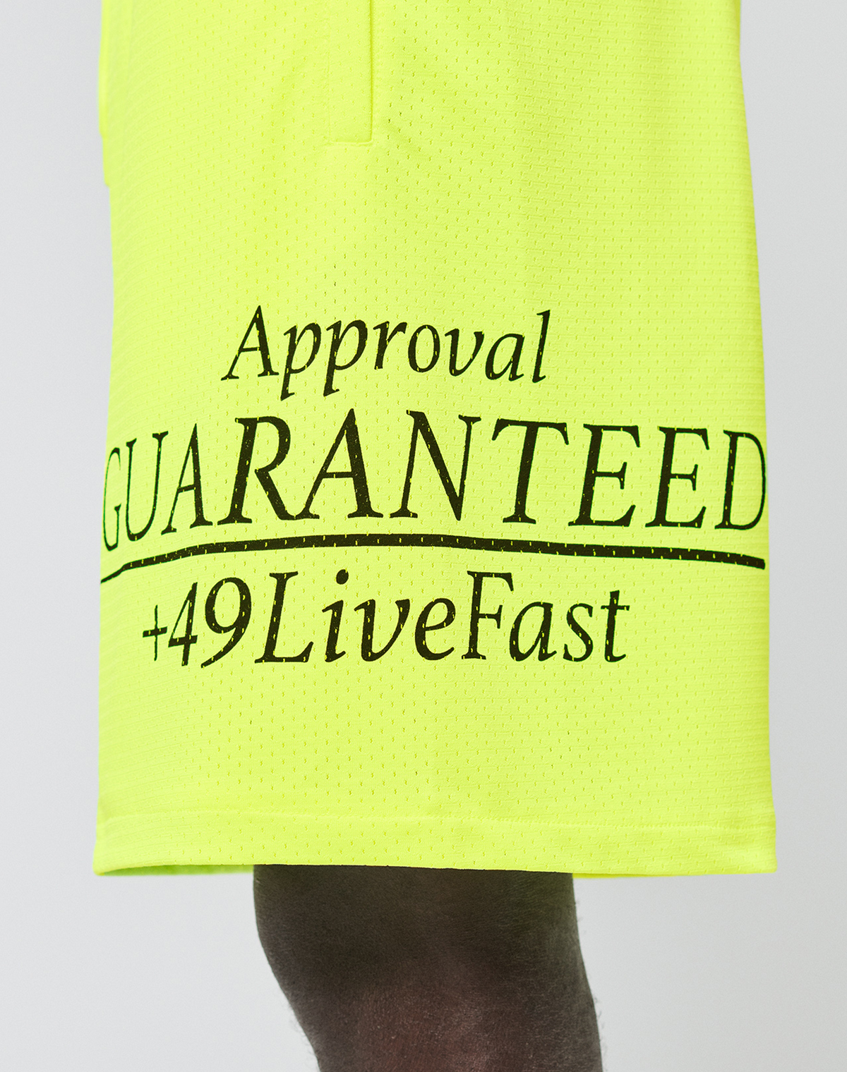 A close-up of LIVE FAST DIE YOUNG’s LFDY Guaranteed Mesh Shorts in bright yellow polyester mesh, featuring black “Approval GUARANTEED +49LiveFast” text. The wide-fit athletic fabric is shown with part of a person’s leg visible.
