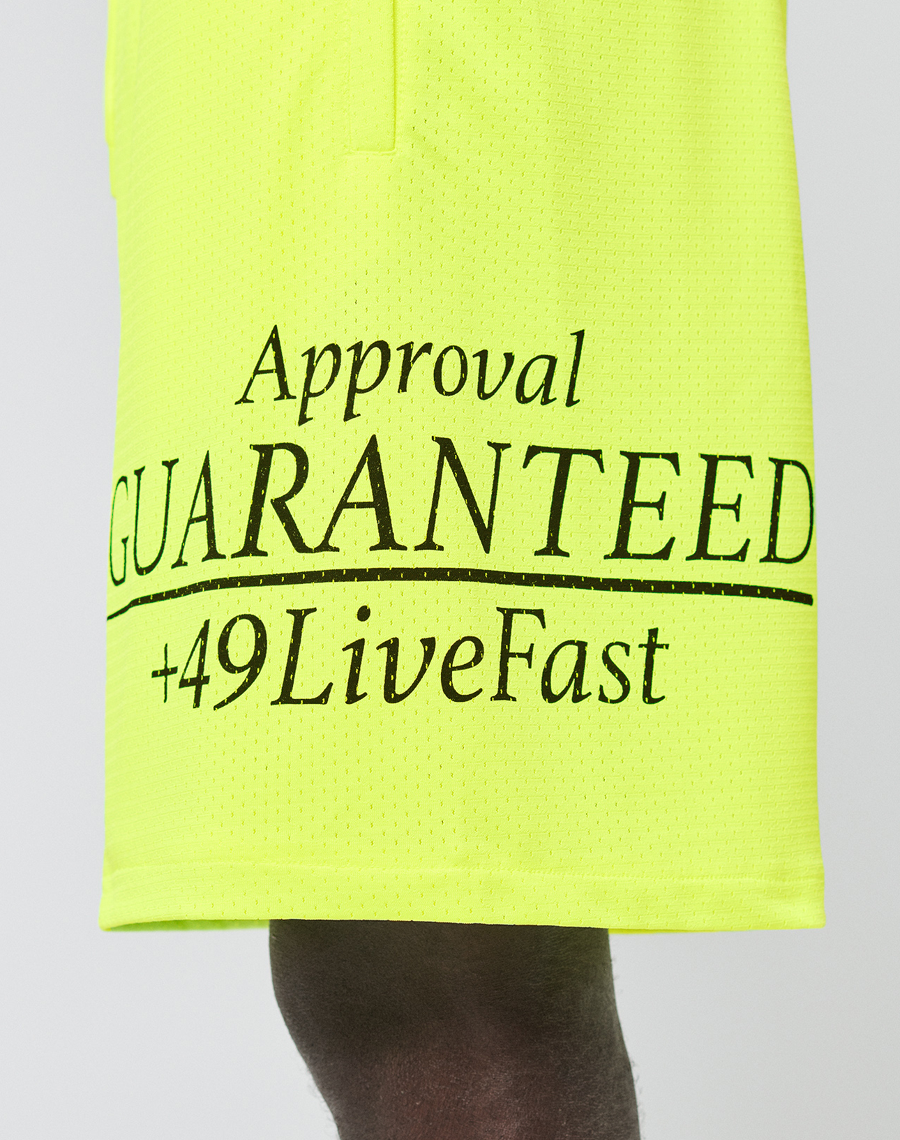 A close-up of LIVE FAST DIE YOUNG’s LFDY Guaranteed Mesh Shorts in bright yellow polyester mesh, featuring black “Approval GUARANTEED +49LiveFast” text. The wide-fit athletic fabric is shown with part of a person’s leg visible.