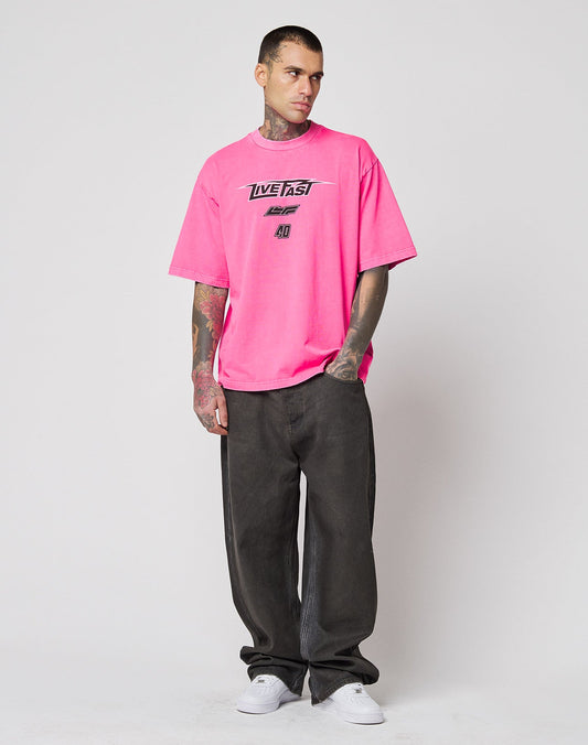 A tattooed man stands against a plain background in the LIVE FAST DIE YOUNG LFDY Triple Racing Tee, an oversize fit with multilogo frontprint, paired with loose dark pants and white sneakers, looking slightly to the side with hands in his pockets.
