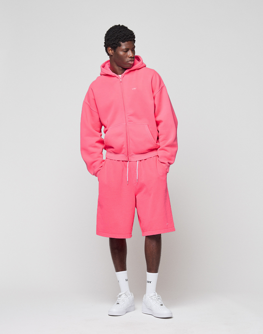A person wears the LIVE FAST DIE YOUNG Basic Hooded Zip in pink with matching shorts, featuring subtle branding, paired with white socks and sneakers. They stand against a plain light gray background, looking to the side with hands in pockets.