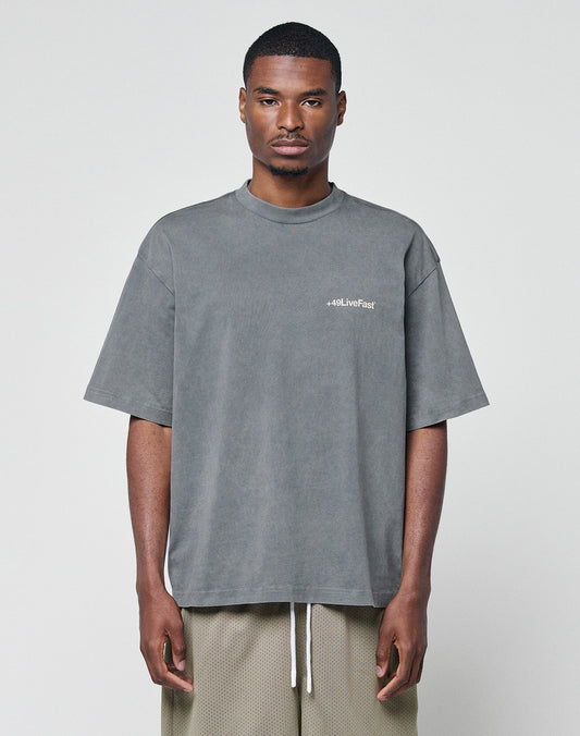 A man faces forward in an oversize fit, faded gray Basic +49 Chest Tee by LIVE FAST DIE YOUNG with a “+49.LiveFast” logo on the left chest and light brown mesh shorts, set against a plain white background. | model