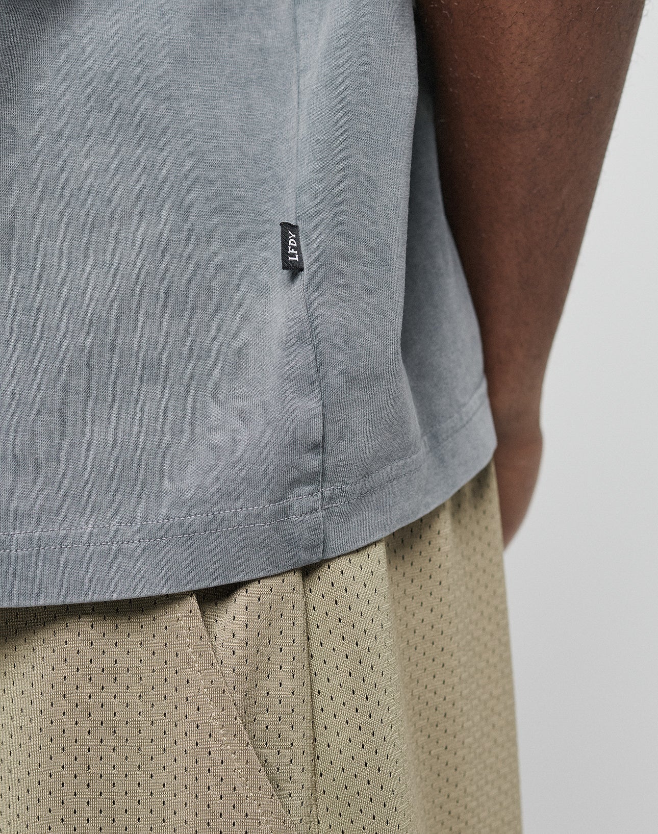 Close-up of a person wearing the LIVE FAST DIE YOUNG Basic +49 Chest Tee in light gray with a small black side tag and beige mesh shorts, focusing on the shirt’s hem and the texture of the shorts.
