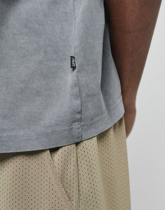 Close-up of a person wearing the LIVE FAST DIE YOUNG Basic +49 Chest Tee in light gray with a small black side tag and beige mesh shorts, focusing on the shirt’s hem and the texture of the shorts.