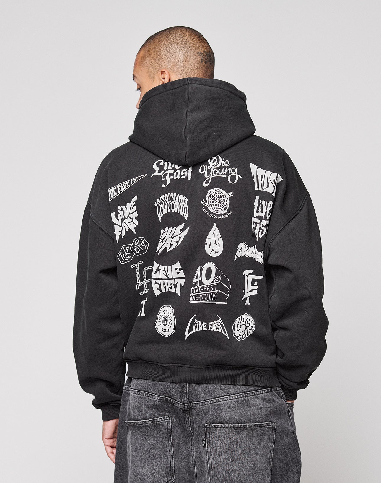 A person in an oversize fit LFDY Logo Hooded Zip by LIVE FAST DIE YOUNG, black with bold white multilogo graphics and phrases like Live Fast, 40 Hours on the back. Made from comfortable Baumwolle, paired with dark jeans and facing away.