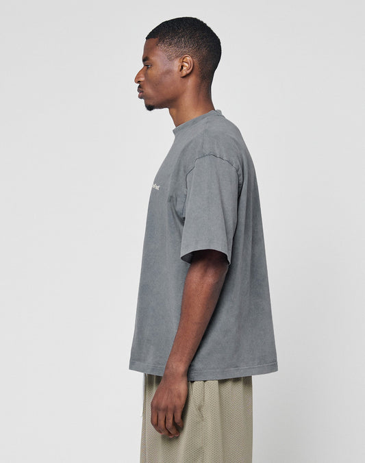 A man stands in profile against a white background, wearing the LIVE FAST DIE YOUNG Basic +49 Chest Tee—an oversize, faded gray 100% Baumwolle T-shirt—paired with light tan mesh shorts.