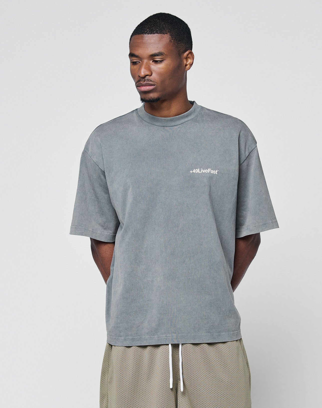 A man stands against a plain background wearing an oversize faded gray LIVE FAST DIE YOUNG Basic +49 Chest Tee with logo on the left chest and beige mesh shorts, looking down with his hands behind his back.