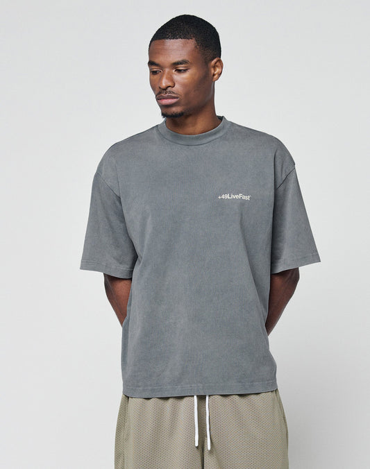 A man stands against a plain background, wearing the LIVE FAST DIE YOUNG Basic +49 Chest Tee in faded gray, 100% Baumwolle, with a subtle left chest "+49LiveFast" logo, paired with light beige mesh shorts with a white drawstring.