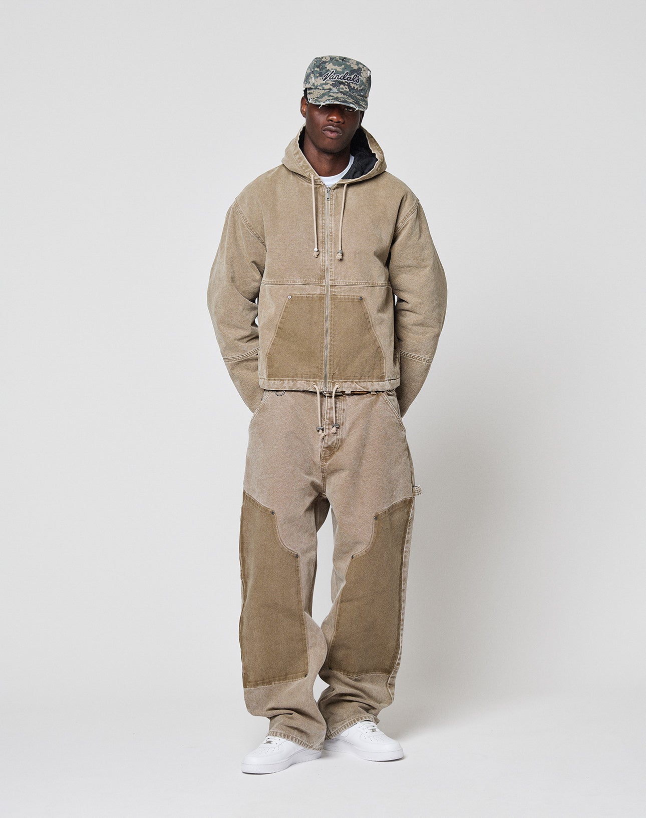 A person in a tan Faded Workwear Jacket by LIVE FAST DIE YOUNG, loose corduroy pants with contrasting knee patches, a camouflage cap, and white sneakers stands against a plain light background.