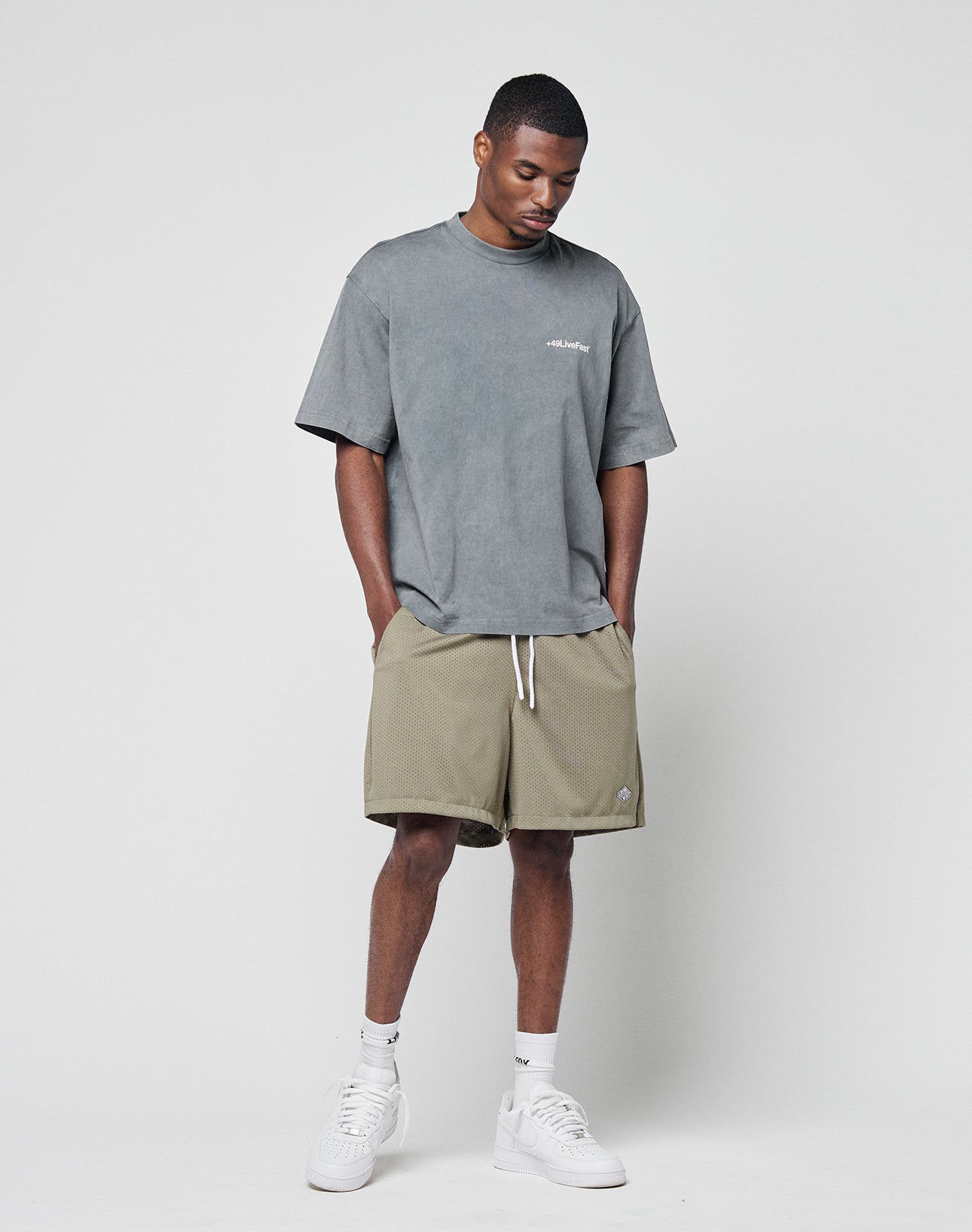 A man stands against a plain background wearing the LIVE FAST DIE YOUNG Basic +49 Chest Tee in grey (oversize fit, 100% Baumwolle), khaki shorts with white drawstrings, white socks, and white sneakers. His head is slightly tilted down with hands in his pockets.