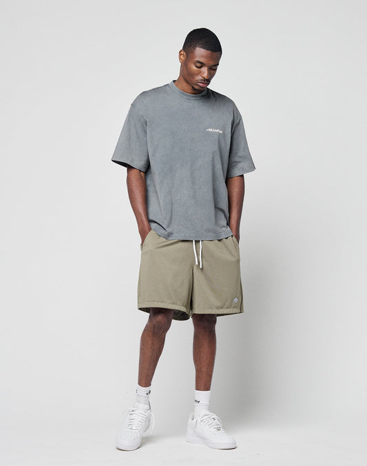 A man stands against a plain background wearing the LIVE FAST DIE YOUNG Basic +49 Chest Tee in grey (oversize fit, 100% Baumwolle), khaki shorts with white drawstrings, white socks, and white sneakers. His head is slightly tilted down with hands in his pockets.