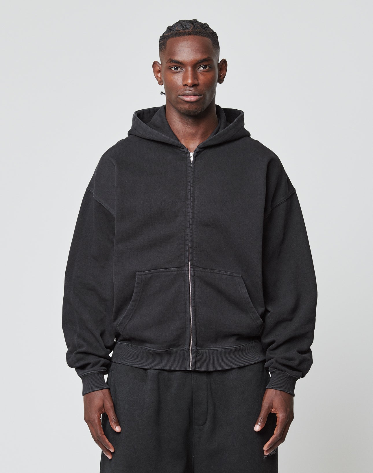 A man faces forward against a light gray background, wearing LIVE FAST DIE YOUNG’s Basic 510 Hooded Zip—an oversize black zip-up hoodie with front pockets, made of 85% Baumwolle—paired with matching black pants.