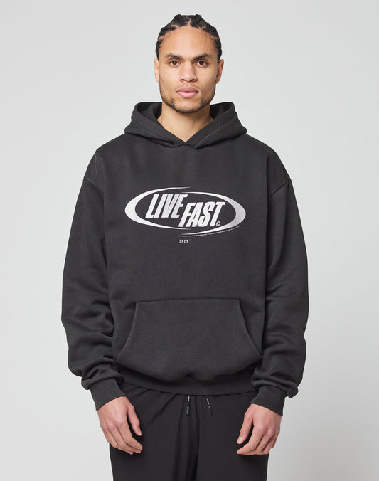 A person in an oversize black LFDY Sphere Hooded by LIVE FAST DIE YOUNG, featuring a bold silver LIVE FAST. oval front print, stands facing forward against a plain light gray background.
