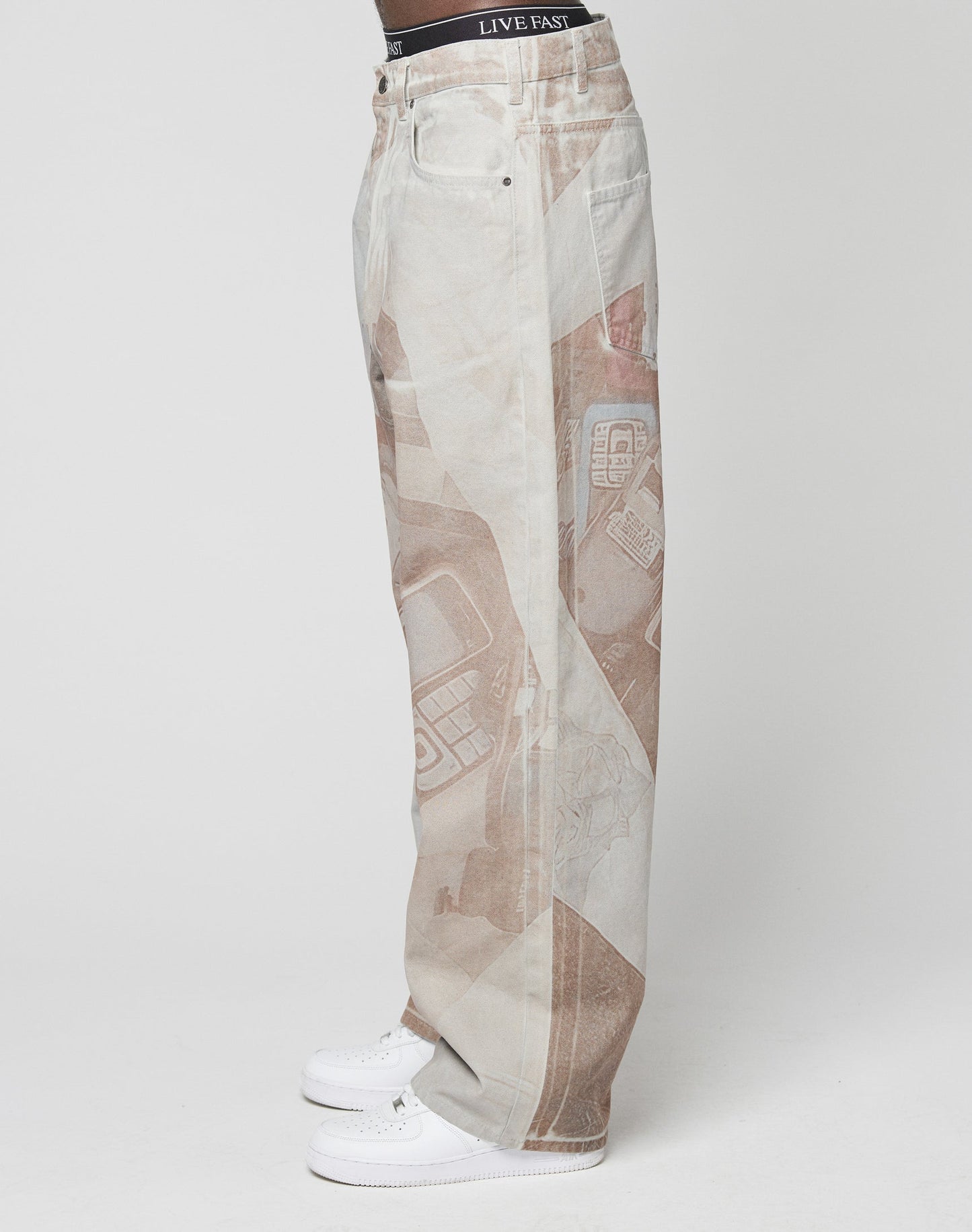 A person stands in profile against a white background, wearing LIVE FAST DIE YOUNG’s LFDY Stolen Phones Denim—baggy, light-wash jeans with abstract brown prints—and white sneakers. The black LIVE FAST waistband peeks above the jeans.