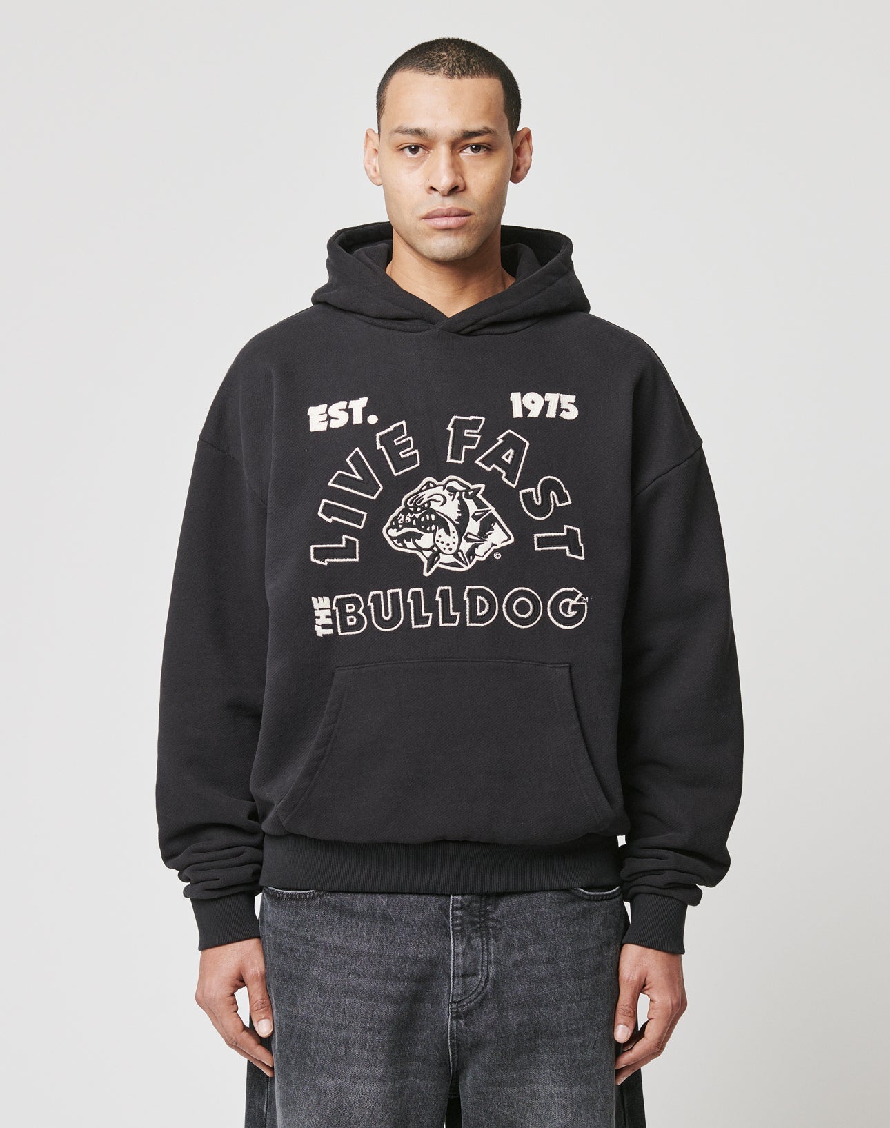 A person in a black LFDY The Bulldog Hooded by LIVE FAST DIE YOUNG, featuring an embroidered EST. 1975 LIVE FAST THE BULLDOG and bulldog graphic, paired with dark jeans, stands against a plain white background.