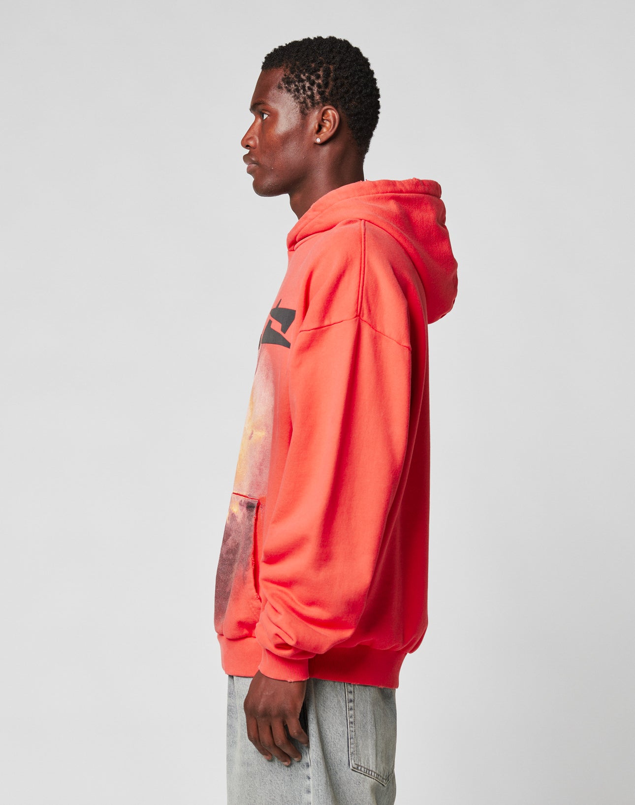 A man in profile wears the LIVE FAST DIE YOUNG LFDY Angel Hooded in bright red with a graphic design and Oversize Fit, paired with light blue jeans featuring a subtle Distress-Effekt, against a plain light gray background.