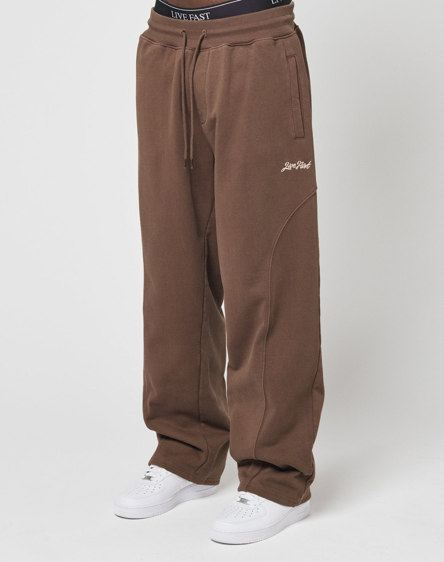 A person wears the LIVE FAST DIE YOUNG Basic Wide Jogger—loose brown sweatpants with a wide elastic waistband and Live Fast on the left thigh—paired with white sneakers and a visible black waistband, against a plain white background.