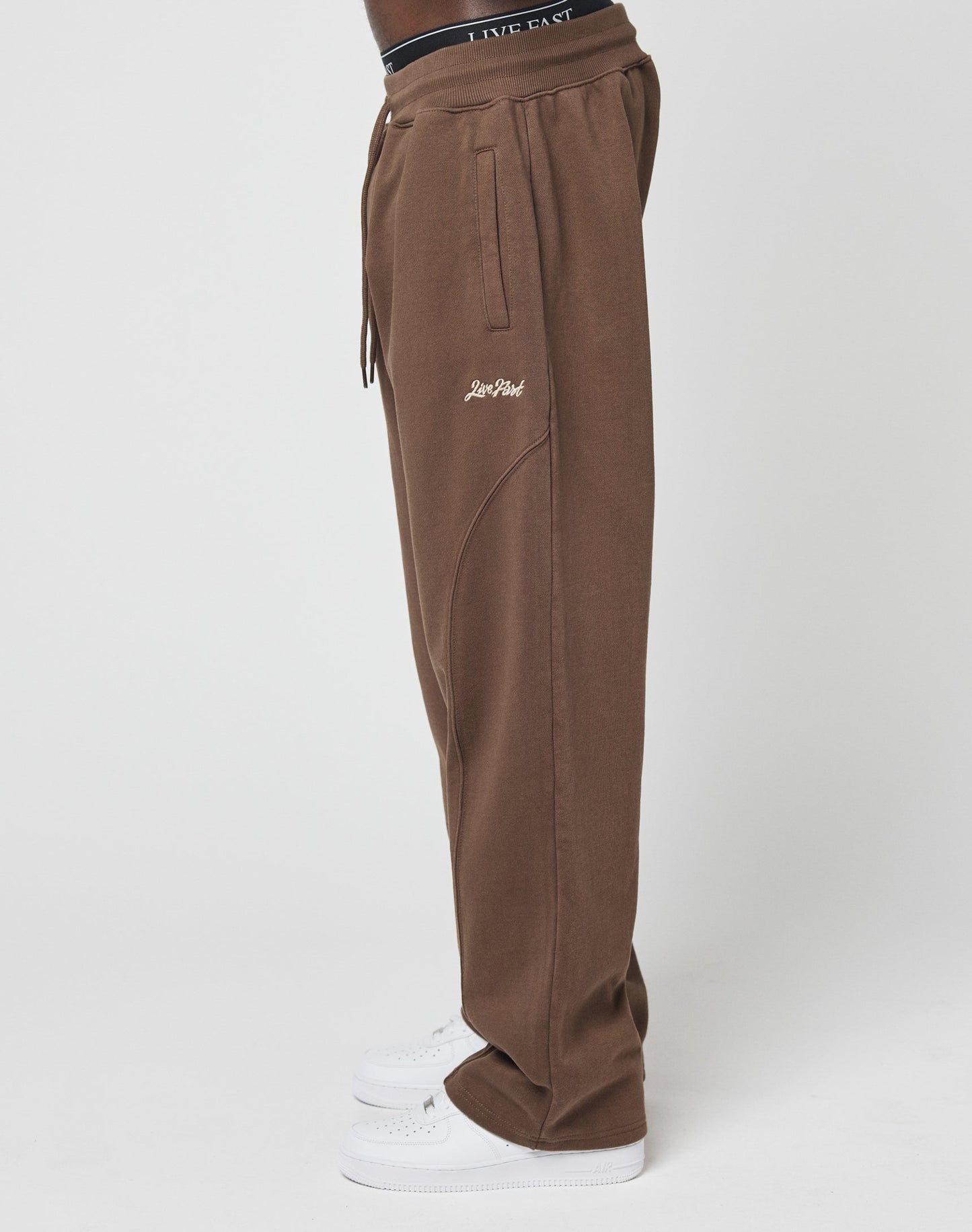 A person in brown Basic Wide Jogger pants by LIVE FAST DIE YOUNG with a wide elastic waistband, side pockets, and white sneakers is shown from the side against a white background. Live Fast is on the waist, Low Heat embroidered on the thigh.