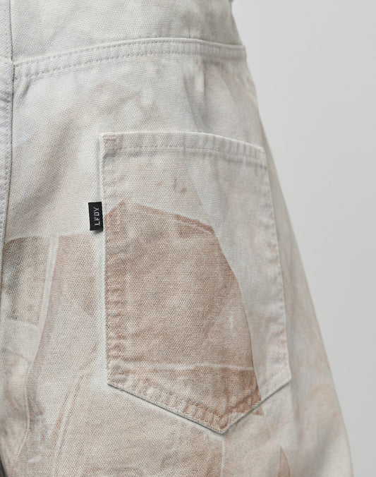 Close-up of the back pocket of LIVE FAST DIE YOUNGs LFDY Stolen Phones Denim jeans. These light-colored, faded denim pants feature a rectangular black JPUY tag, subtle brown abstract all-over print, visible stitching, and are 100% cotton.