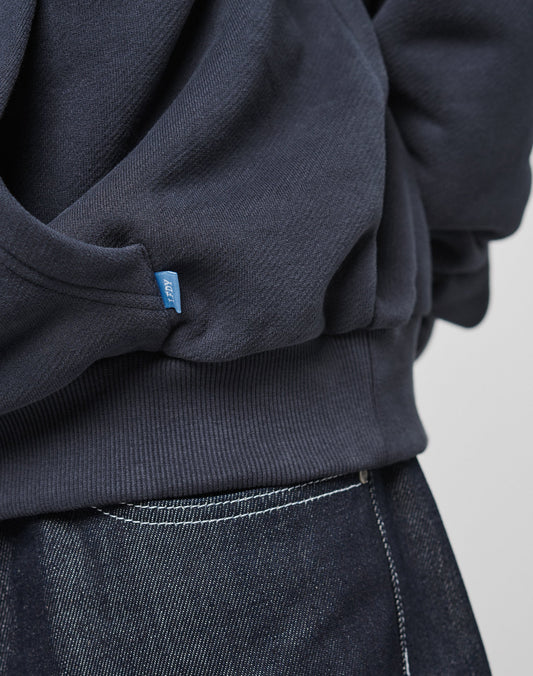 Close-up of a person wearing the LIVE FAST DIE YOUNG LFDY Script Hooded sweatshirt in dark navy waffle fabric with a blue tag, paired with dark denim jeans with white stitching. Only the lower oversize-fit sweatshirt and jean waistband are visible.