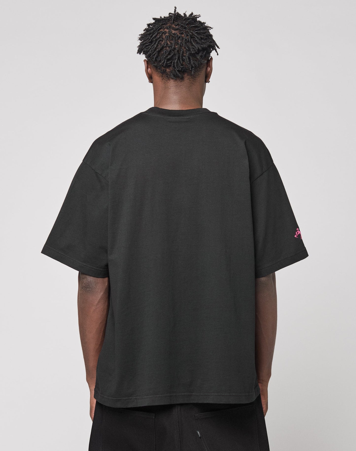 A person with short, textured hair stands with their back to the camera, wearing the LIVE FAST DIE YOUNG Live Fast Stallion Tee in black and black pants against a plain light background.