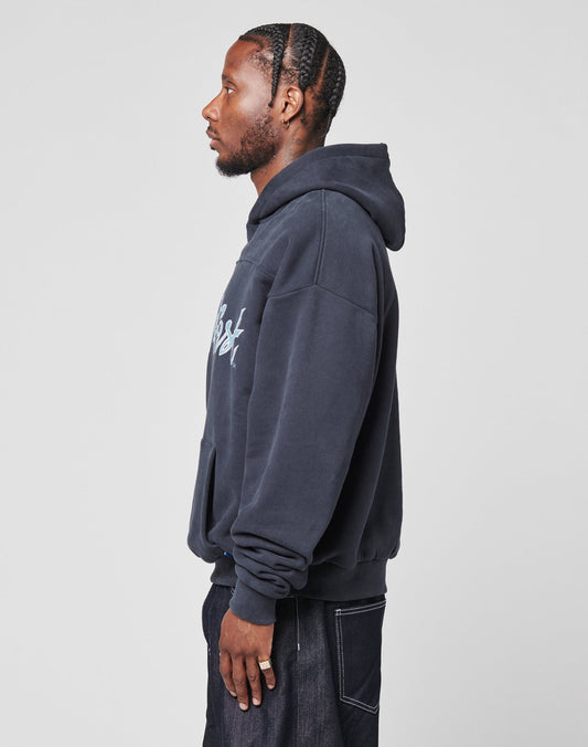 A man with braided hair wears the LIVE FAST DIE YOUNG LFDY Script Hooded in an oversize fit with dark jeans, standing in profile against a plain light gray background.
