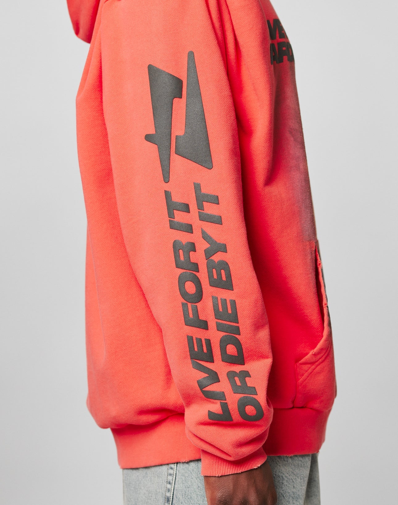A person in a LIVE FAST DIE YOUNG LFDY Angel Hooded red hoodie with black graphic and “LIVE FOR IT OR DIE BY IT” on the sleeve, made of 100% Baumwolle, paired with light blue jeans in a stylish side profile.