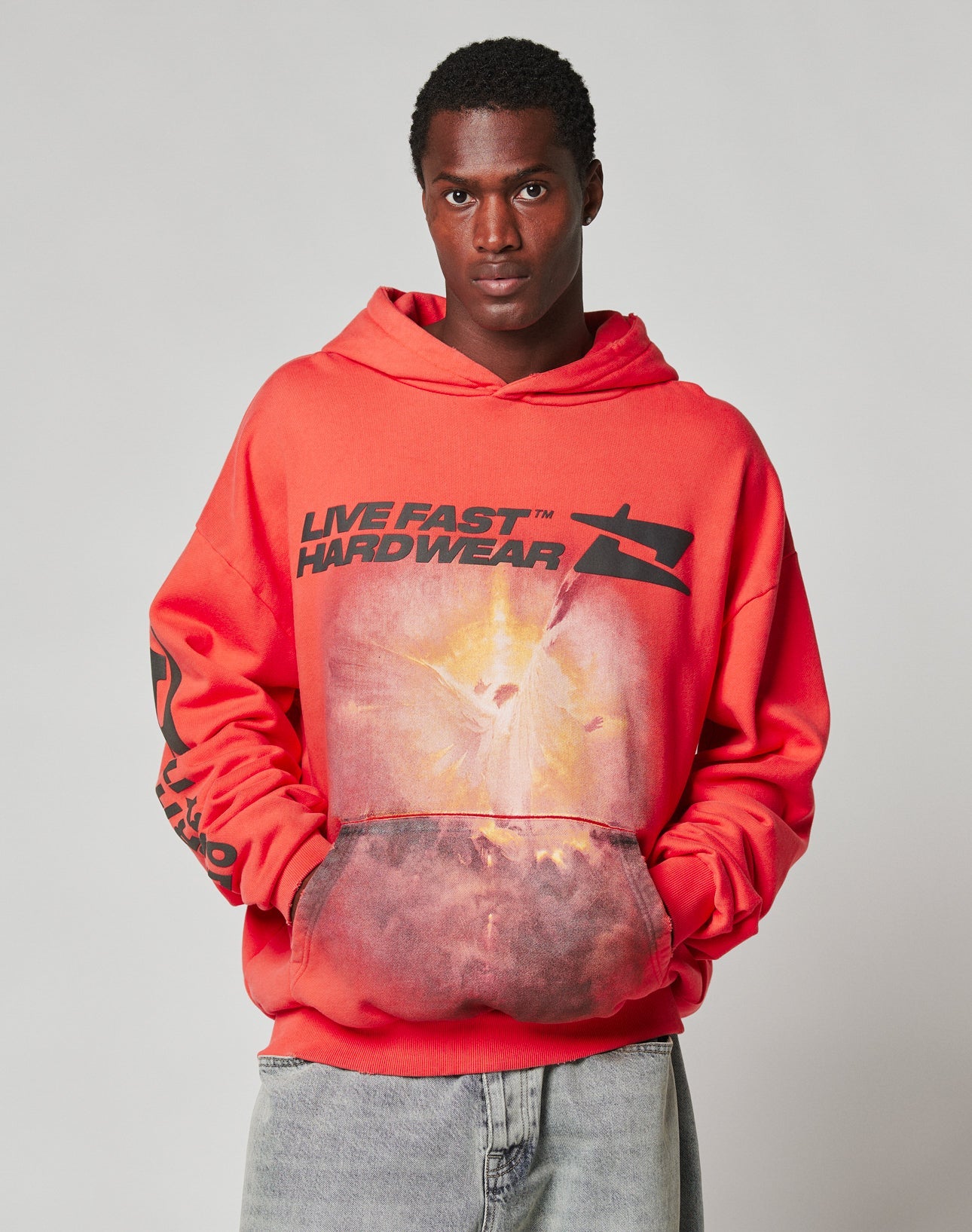 A young man wears the LFDY Angel Hooded by LIVE FAST DIE YOUNG, an oversized bright red 100% Baumwolle hoodie with bold black LIVE FAST HARDWEAR text and an angel graphic, standing against a plain gray background with hands in pockets.