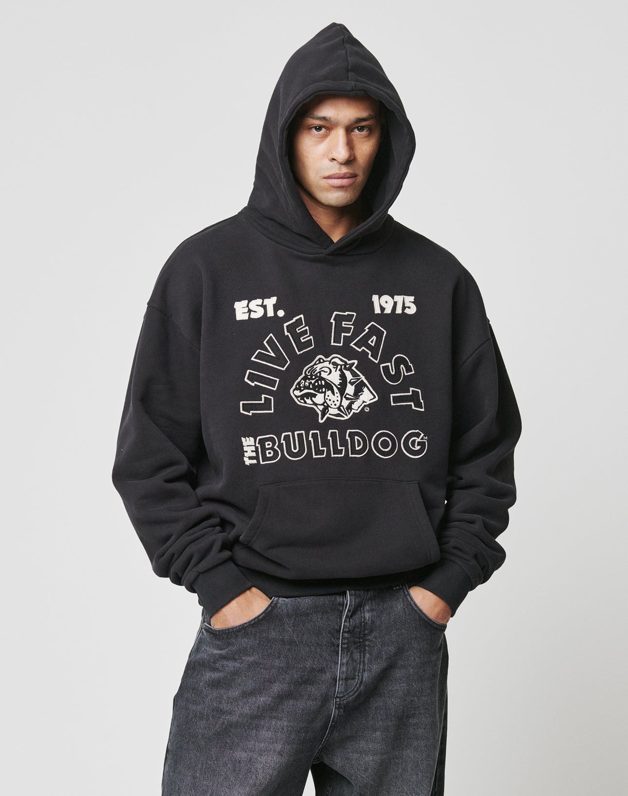 A person in an oversize black LFDY The Bulldog Hooded by LIVE FAST DIE YOUNG, with hood up and hands in pockets, features a bulldog graphic and embroidered LIVE FAST, EST. 1975, and BULLDOG on the front against a plain background.