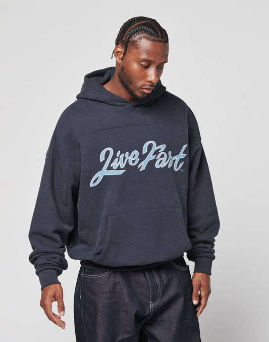 A man with braided hair wears the LIVE FAST DIE YOUNG LFDY Script Hooded in oversize navy blue, featuring Live Part in large script on the front, paired with distressed dark jeans, standing against a plain light background.
