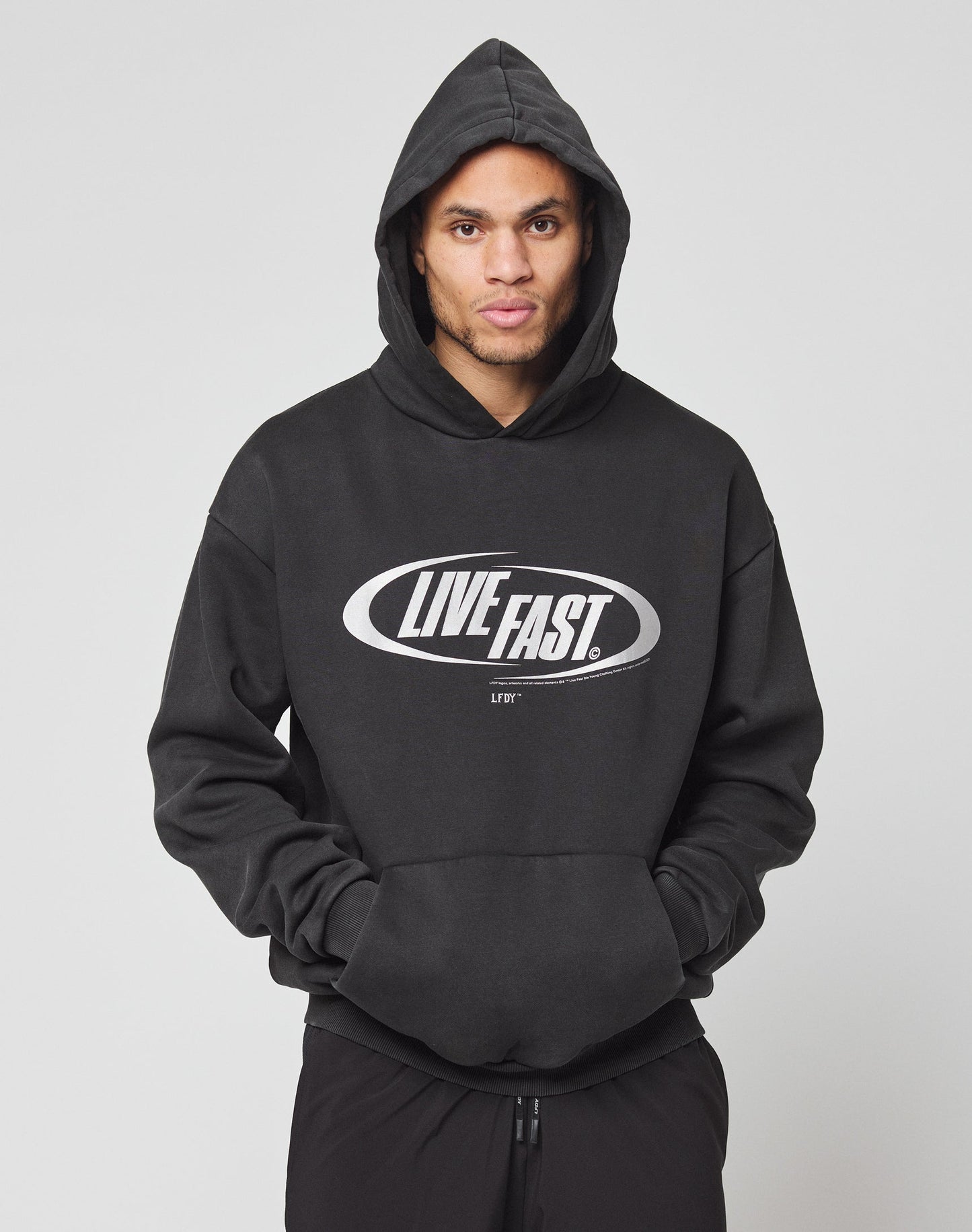 Someone stands against a plain light gray background wearing the LFDY Sphere Hooded by LIVE FAST DIE YOUNG—an oversized black hoodie featuring a silver LIVE FAST. front print, with the hood up.