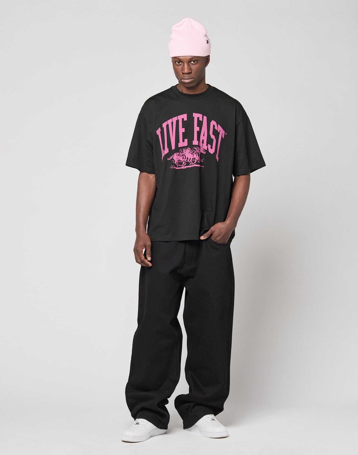 A person wears the LIVE FAST DIE YOUNG Live Fast Stallion Tee, an oversized black T-shirt with bold pink LIVE FAST front print, paired with a pink beanie, wide black pants, and white sneakers. Made from 100% Baumwolle.