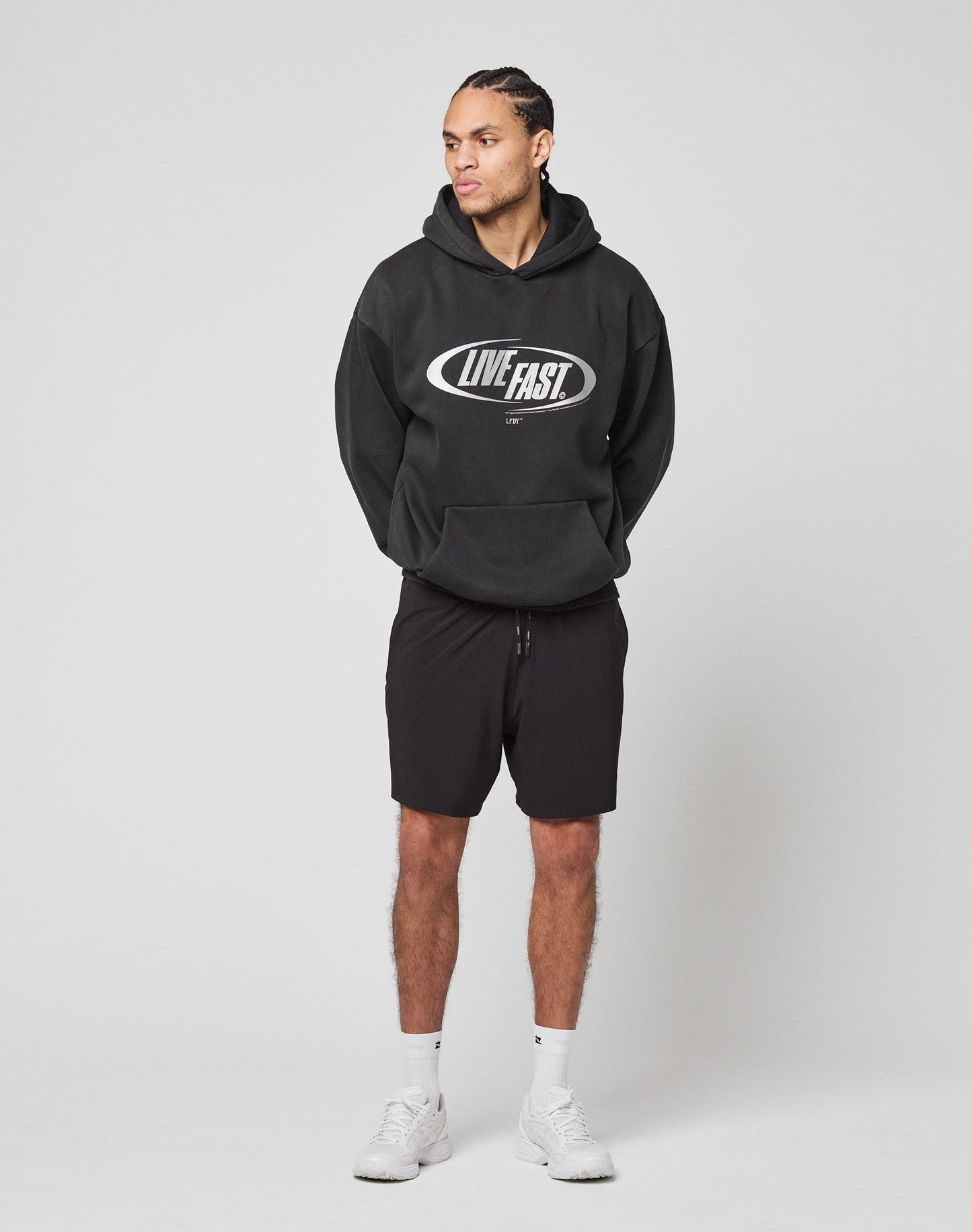 A person poses against a plain background wearing the black LFDY Sphere Hooded by LIVE FAST DIE YOUNG with silver front print in an oversized fit, paired with black shorts, white socks, and white athletic shoes, looking slightly to the side.