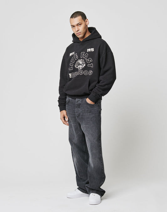 A man wearing the LFDY The Bulldog Hooded by LIVE FAST DIE YOUNG, paired with loose black jeans and white sneakers, stands against a plain background looking to the side in a relaxed pose.
