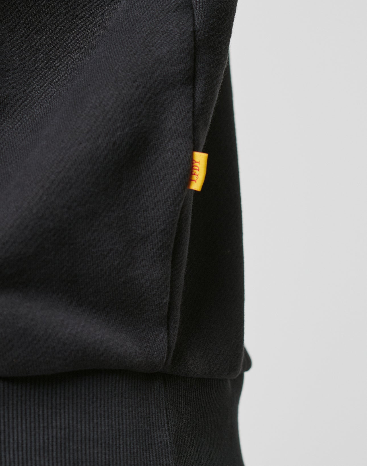 Close-up of LFDY The Bulldog Hooded by LIVE FAST DIE YOUNG, showing black fabric with a small yellow tag stitched into the seam. The fabric’s quality and detailed embroidery are visible against a plain, light background.