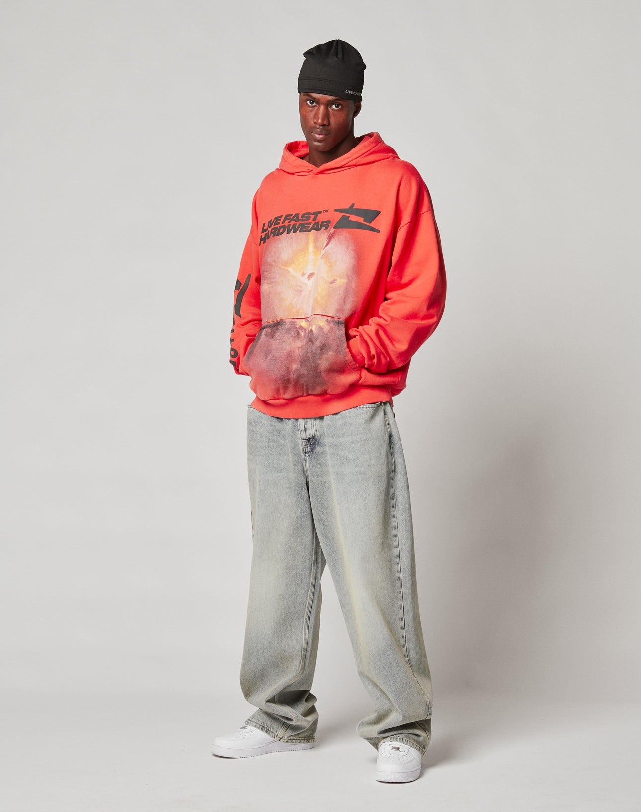 A person stands against a plain background wearing the red LFDY Angel Hooded by LIVE FAST DIE YOUNG, paired with baggy light-wash oversize jeans, white sneakers, and a black beanie, looking confidently at the camera.