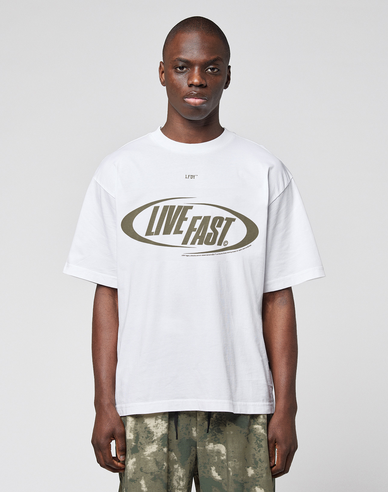 A person wears the LFDY Sphere Tee by LIVE FAST DIE YOUNG, a white oversize cotton T-shirt with "LIVE FAST" in bold inside an oval, featuring front and back prints, paired with camouflage pants. | model