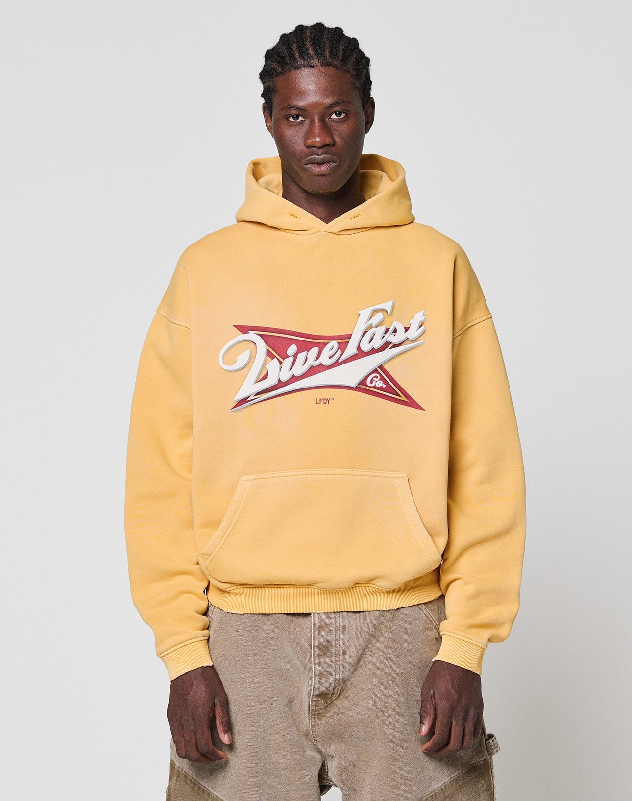 A person wears an oversize yellow LFDY Corp. Hooded by LIVE FAST DIE YOUNG, featuring a Drive Fast spray-effect front print, with tan pants, standing against a plain light gray background and looking at the camera neutrally.