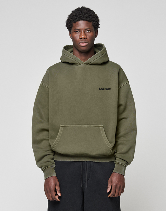 A person wears the LIVE FAST DIE YOUNG Basic 360 Hooded in olive green with topstitching and "LiveFast" on the chest, styled with black pants and standing against a plain light gray background, looking at the camera. The hoodie has an oversize fit.