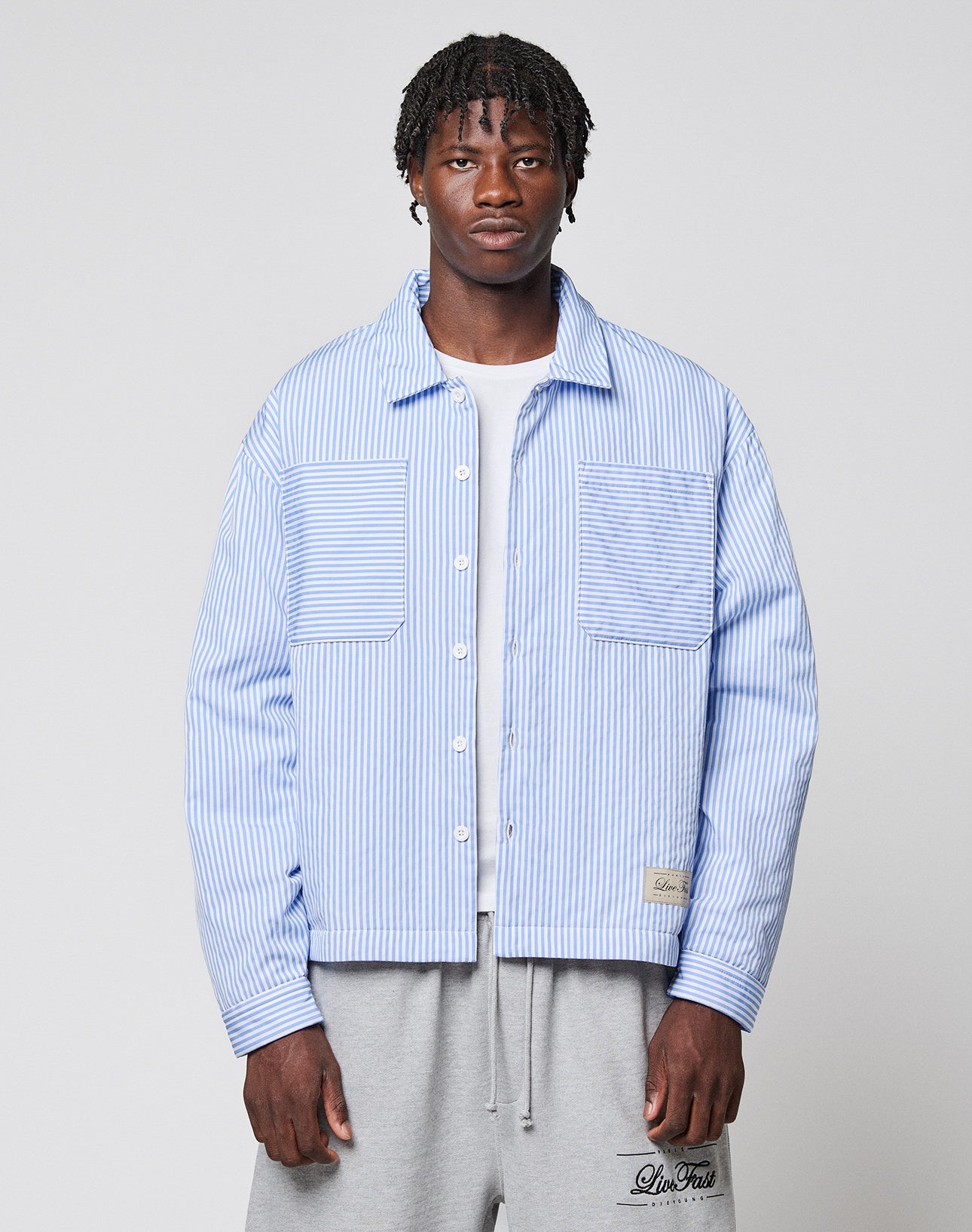 A man with short, styled hair and a neutral expression wears the LIVE FAST DIE YOUNG LFDY Striped Padded Shirt over a white t-shirt and gray sweatpants, standing against a plain background.