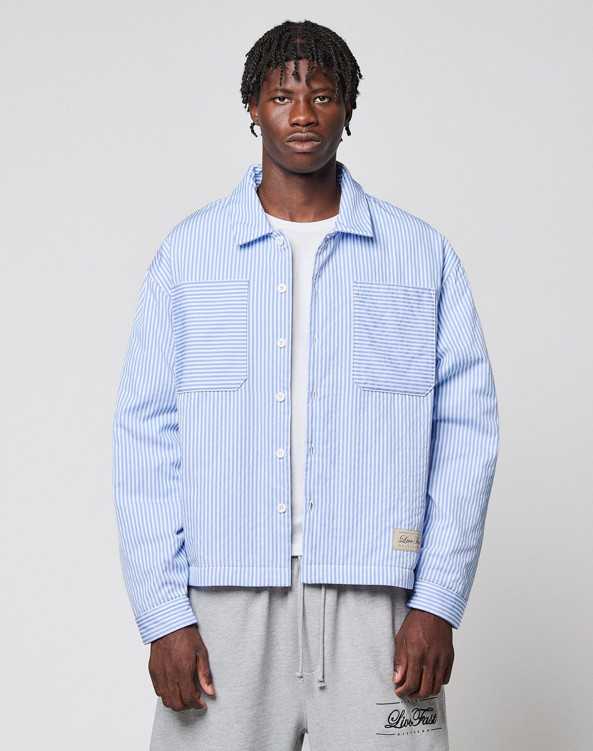 A man with short, styled hair and a neutral expression wears the LIVE FAST DIE YOUNG LFDY Striped Padded Shirt over a white t-shirt and gray sweatpants, standing against a plain background. | model