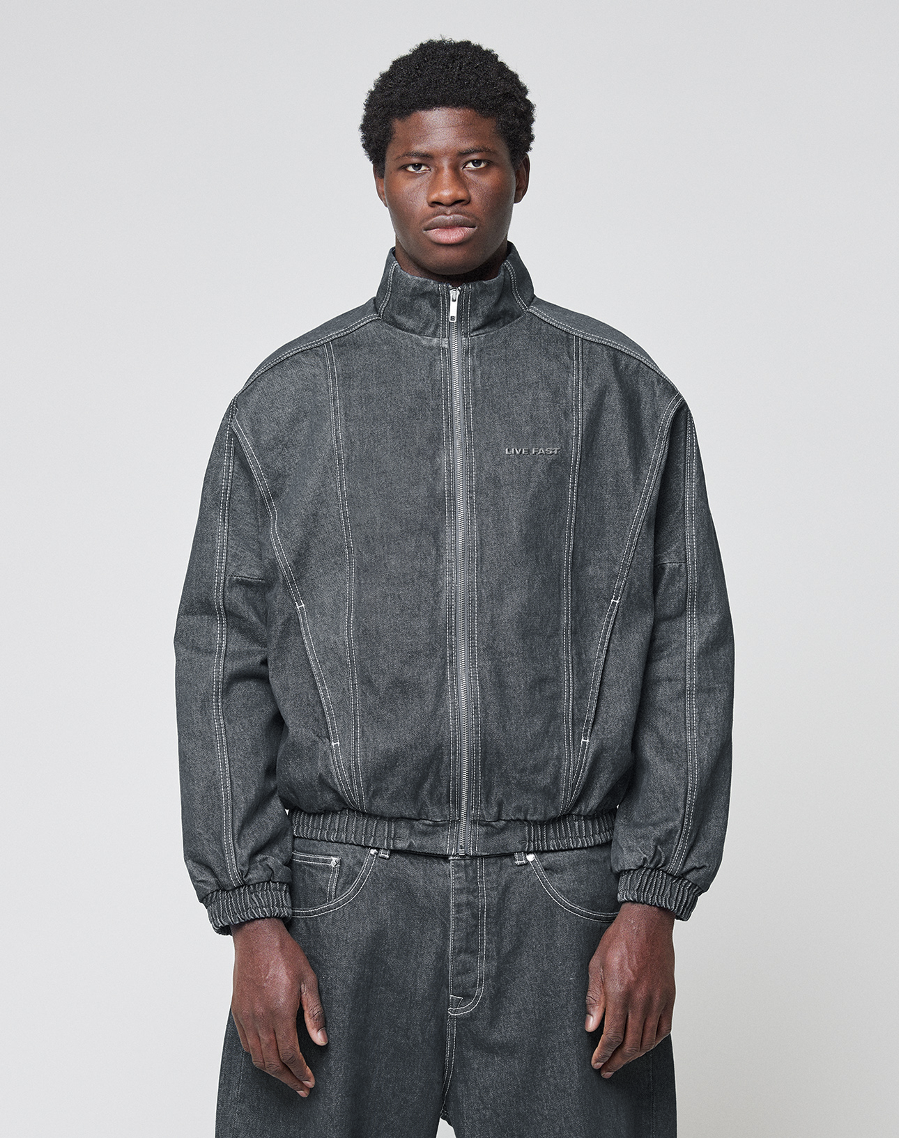 A man faces forward in a dark gray LFDY Denim Trackjacket by LIVE FAST DIE YOUNG, with visible seams, subtle chest text, and paired baggy pants. The 100% cotton jacket features a stand-up collar and is set against a plain light background. | model